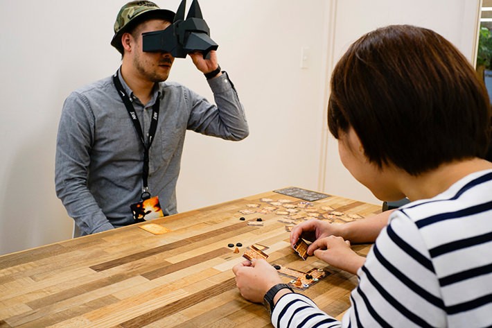 向かって左の男性がVRでピラミッドを覗き、自分には何が見えているのかを他のプレーヤーに伝えています